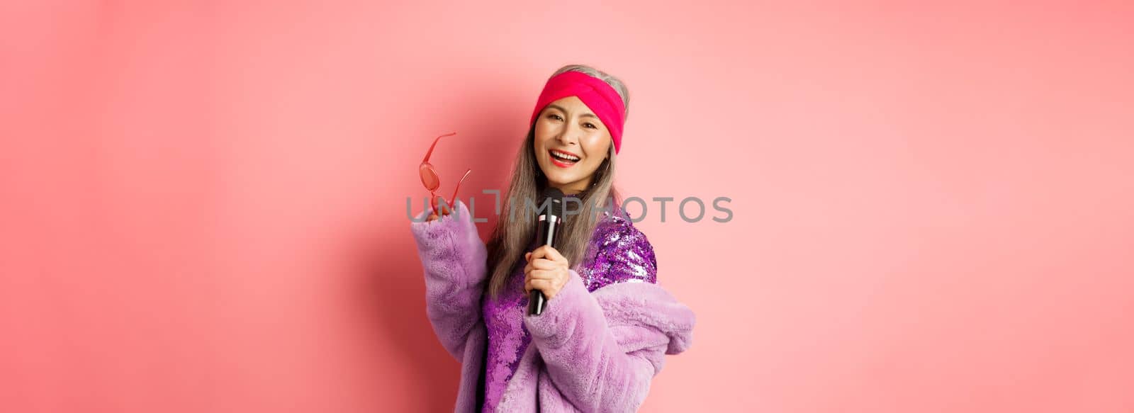 Gorgeous asian senior woman singing karaoke in mic, perform song and looking happy, standing over pink background by Benzoix