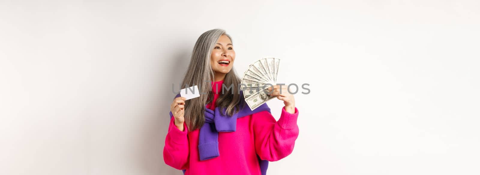 Shopping concept. Stylish asian senior woman showing money dollars and plastic credit card, looking dreamy aside, thinking about buying, white background.