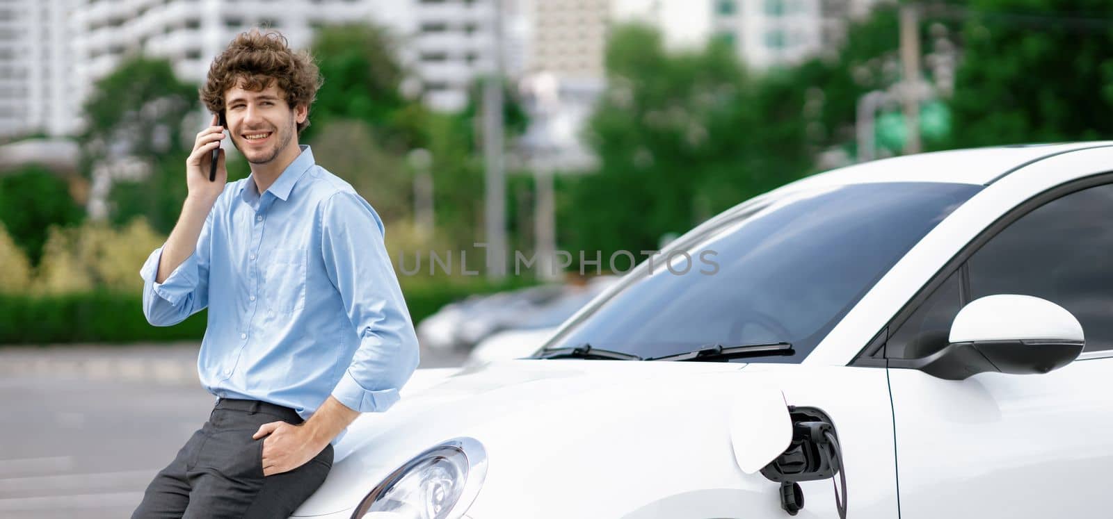 Progressive businessman talking on the phone, leaning on electric car recharging with public EV charging station, apartment condo residential building on the background as green city lifestyle.