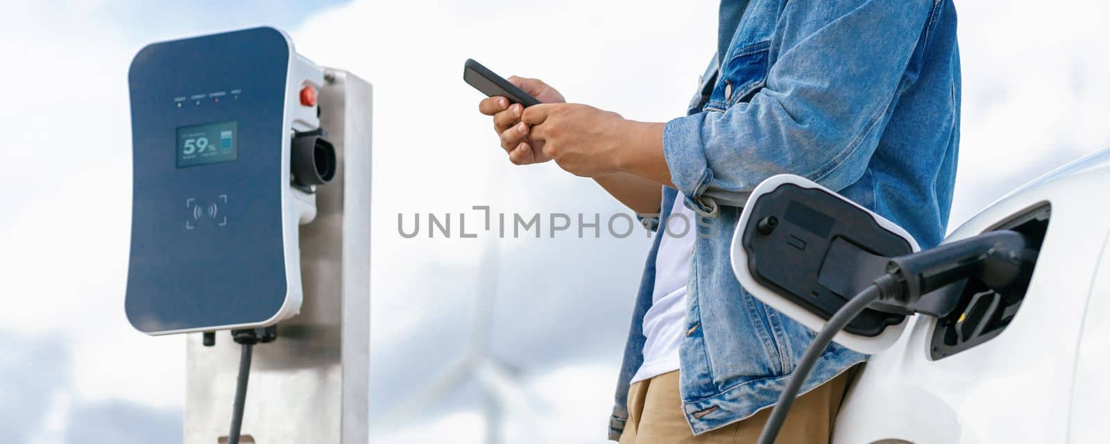 Progressive man with his electric car, EV car recharging energy from charging station on green field with wind turbine as concept of future sustainable energy. Electric vehicle with energy generator.