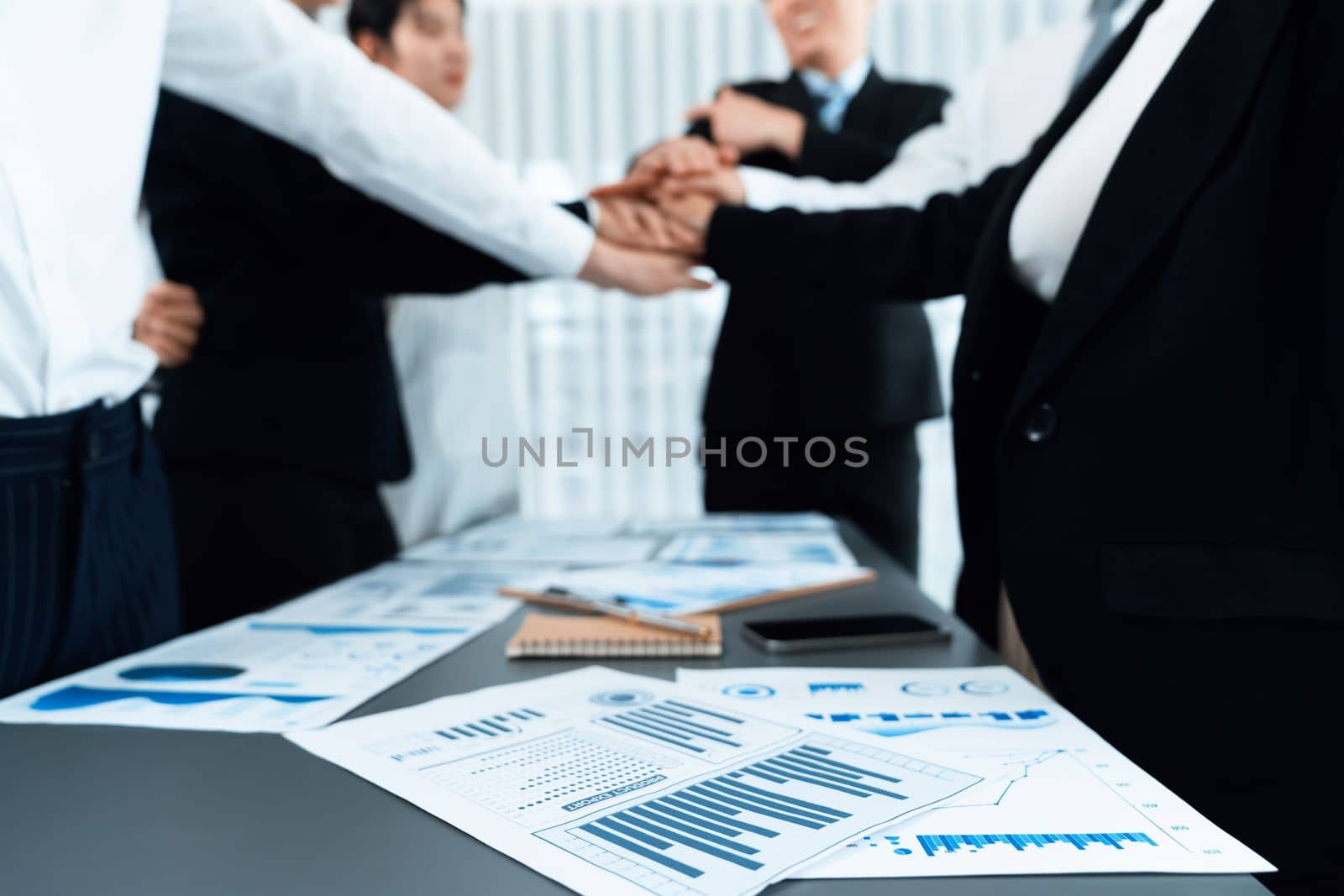 Focus financial report with statistic charts analyzed by BI papers on meeting table at conference room with blur business colleague people put hand stack together in background as harmony concept.