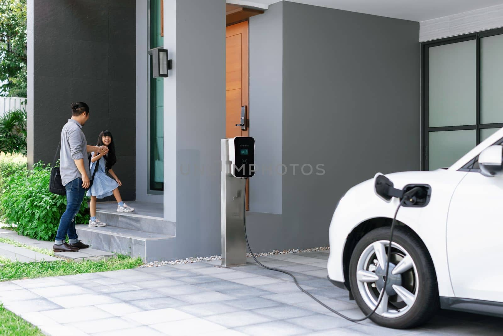 Progressive father and daughter plugs EV charger from home charging station to electric vehicle. Future eco-friendly car with EV cars powered by renewable source of clean energy.