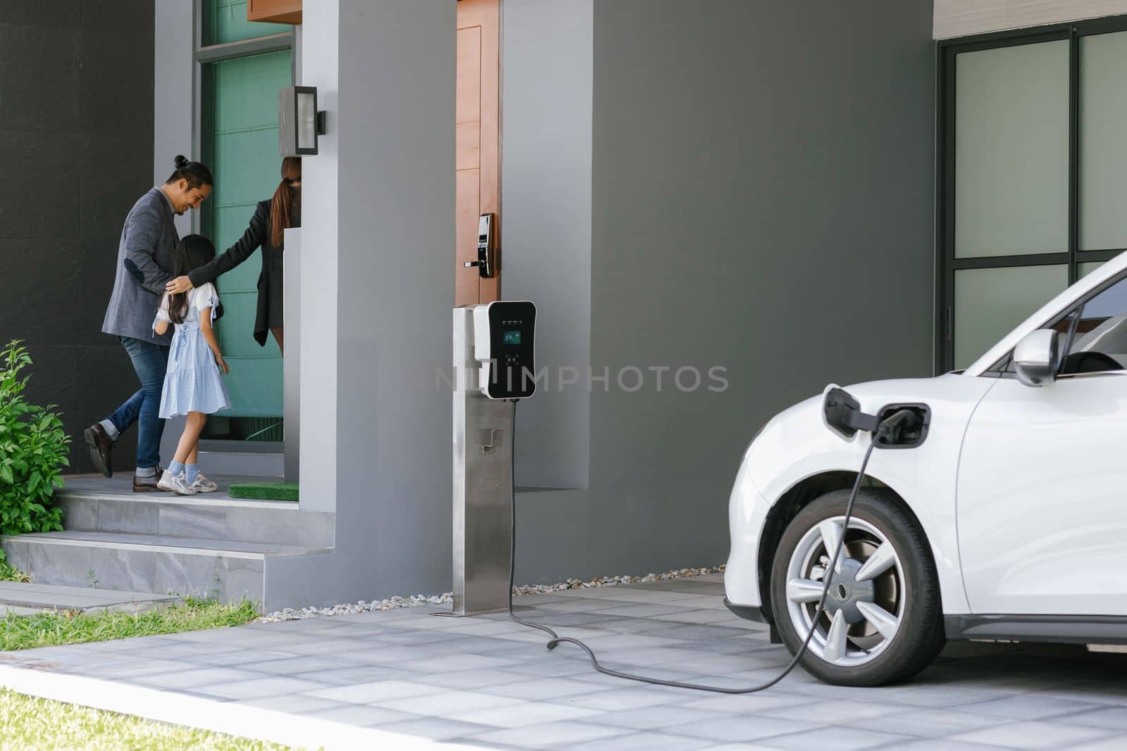 Progressive young parents and daughter with electric vehicle and home charging station. Green and clean energy from electric vehicles for healthy environment. Eco power from renewable source at home.