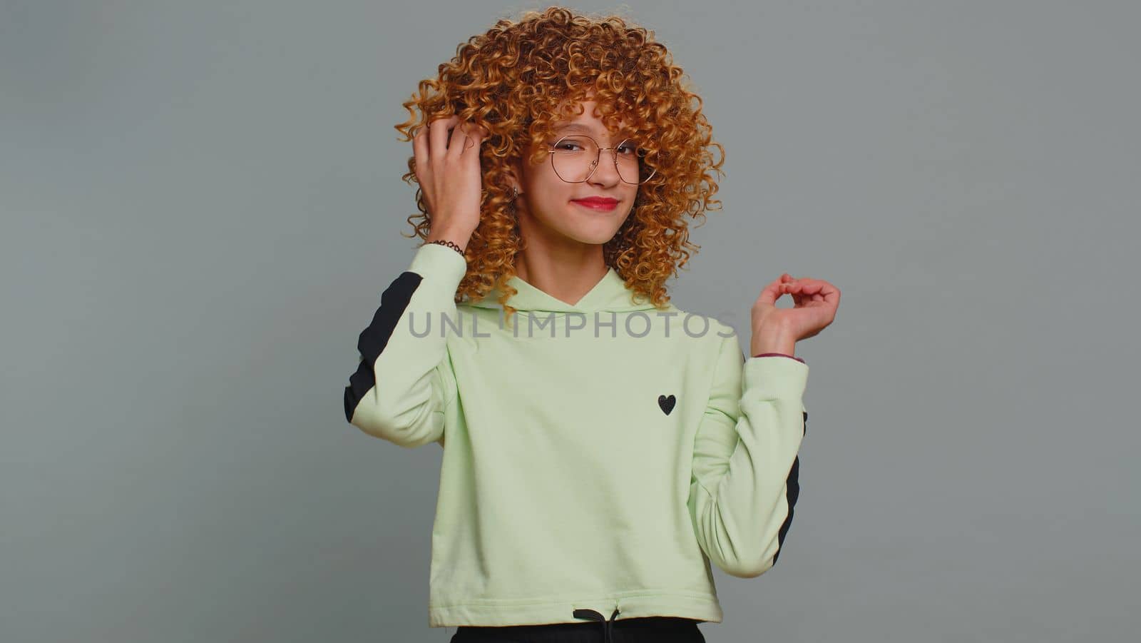 Cheerful lovely young child girl kid in glasses smiling looking at camera. Funny female teenager children schoolgirl with curly frizzy hair isolated portrait alone on studio gray background. Childhood