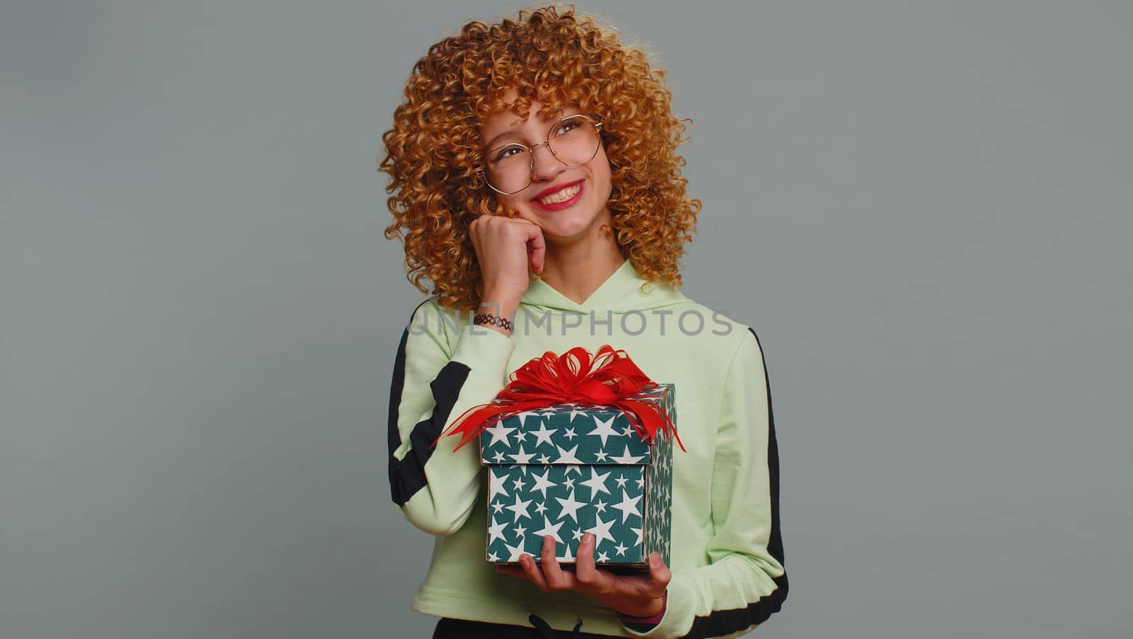 Happy teenager school girl receive, opening gift box and smiling joyfully, looking amazed satisfied with nice present unexpected birthday surprise. Curly haired child kid isolated on gray background