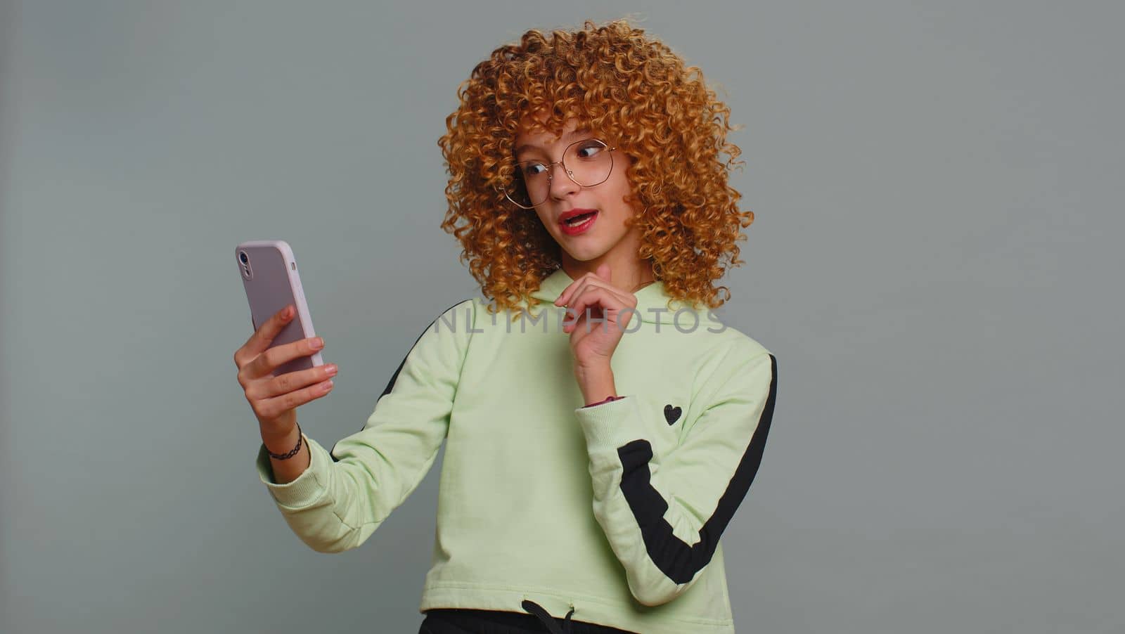 Young child girl kid blogger taking selfie on smartphone holding in hand, communicating video call talking online with subscribers. Teenager children with curly hair isolated on studio gray background