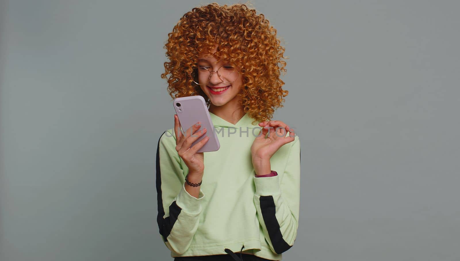 Young child girl kid with curly hair use mobile smartphone typing browsing say Wow yes found out great big win good news celebrate doing winner gesture. Teenager children on studio gray background