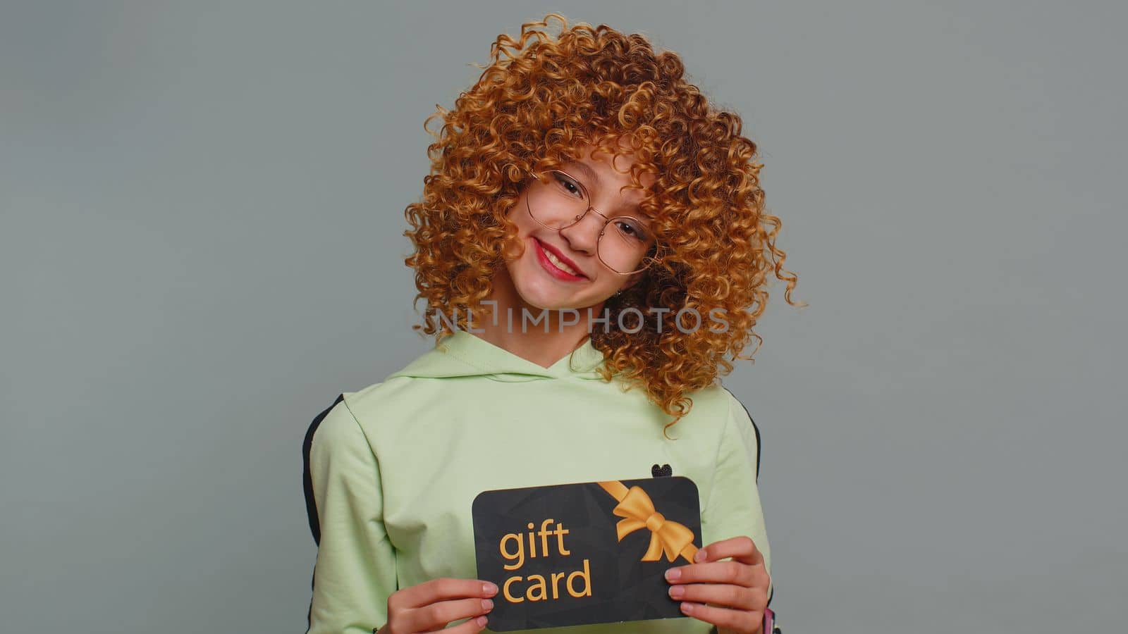 Teenager school girl showing pointing on gift discount certificate voucher coupon for store holidays sale. Child kid with shopping christmas surprise card isolated alone on gray studio background