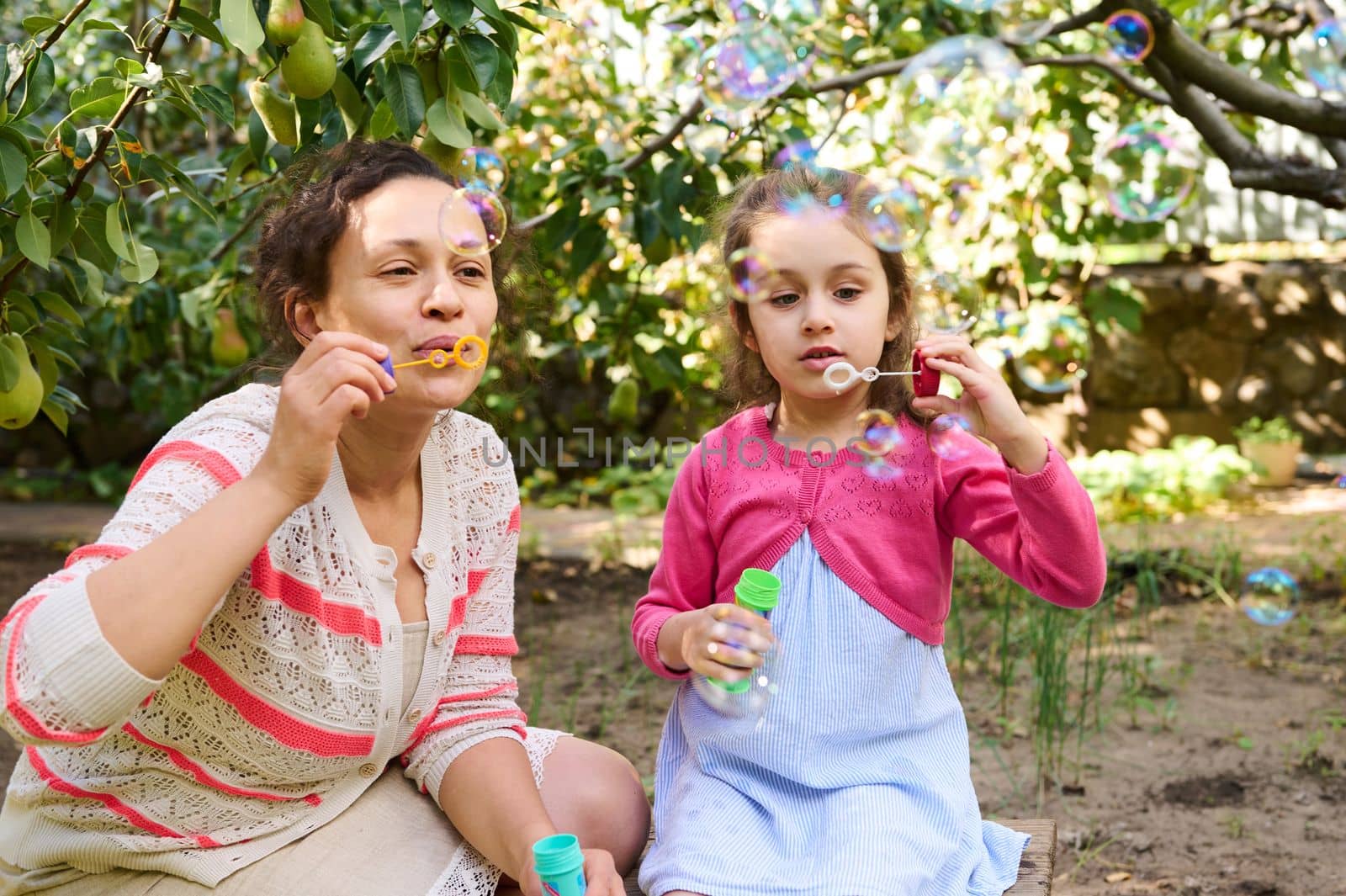 Happy loving mother and daughter blowing soap bubbles, having fun together in an eco garden, sitting under a tree with ripening pears in the countryside. Weekend with family. mom and baby together