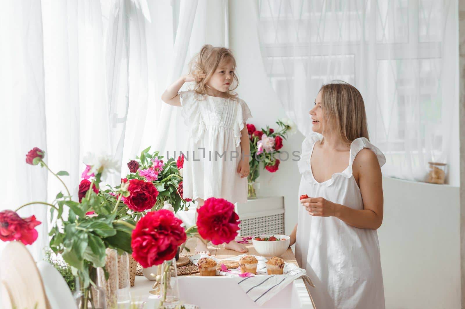 A little blonde girl with her mom on a kitchen countertop decorated with peonies. The concept of the relationship between mother and daughter. Spring atmosphere.