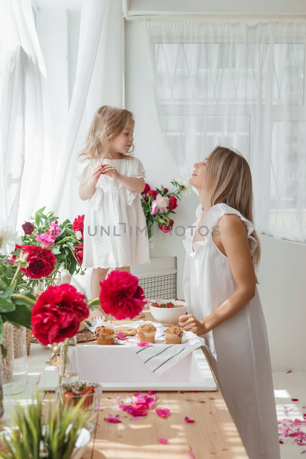 A little blonde girl with her mom on a kitchen countertop decorated with peonies. The concept of the relationship between mother and daughter. Spring atmosphere.