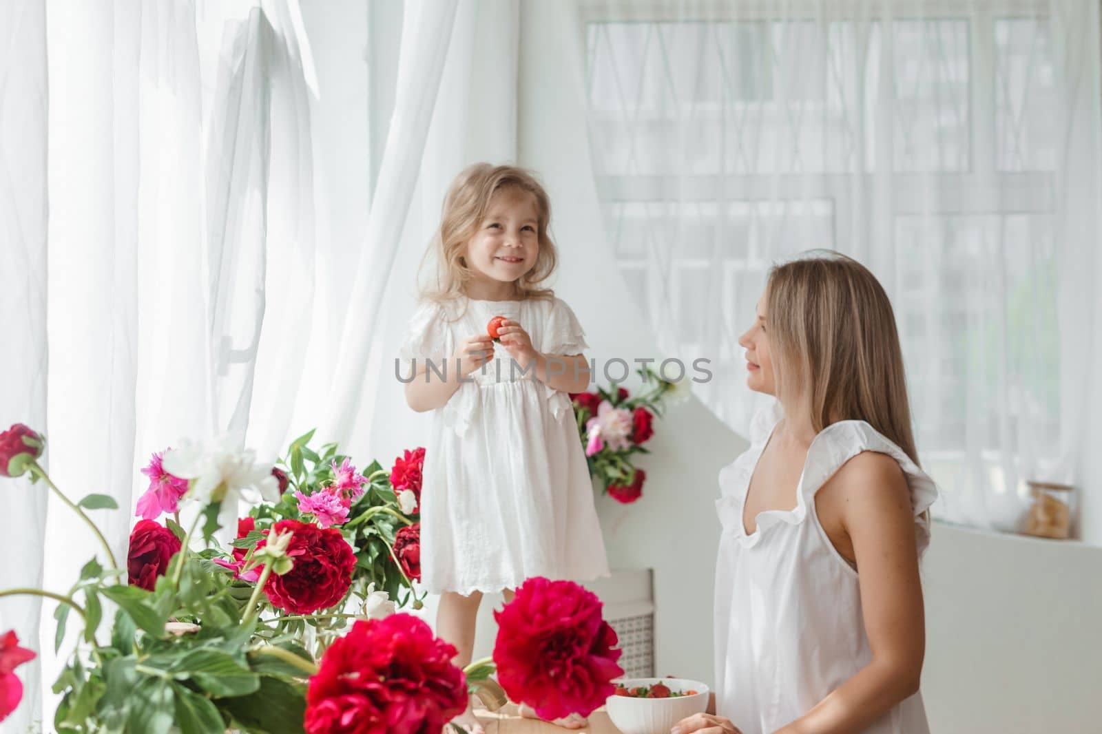 A little blonde girl with her mom on a kitchen countertop decorated with peonies. The concept of the relationship between mother and daughter. Spring atmosphere.