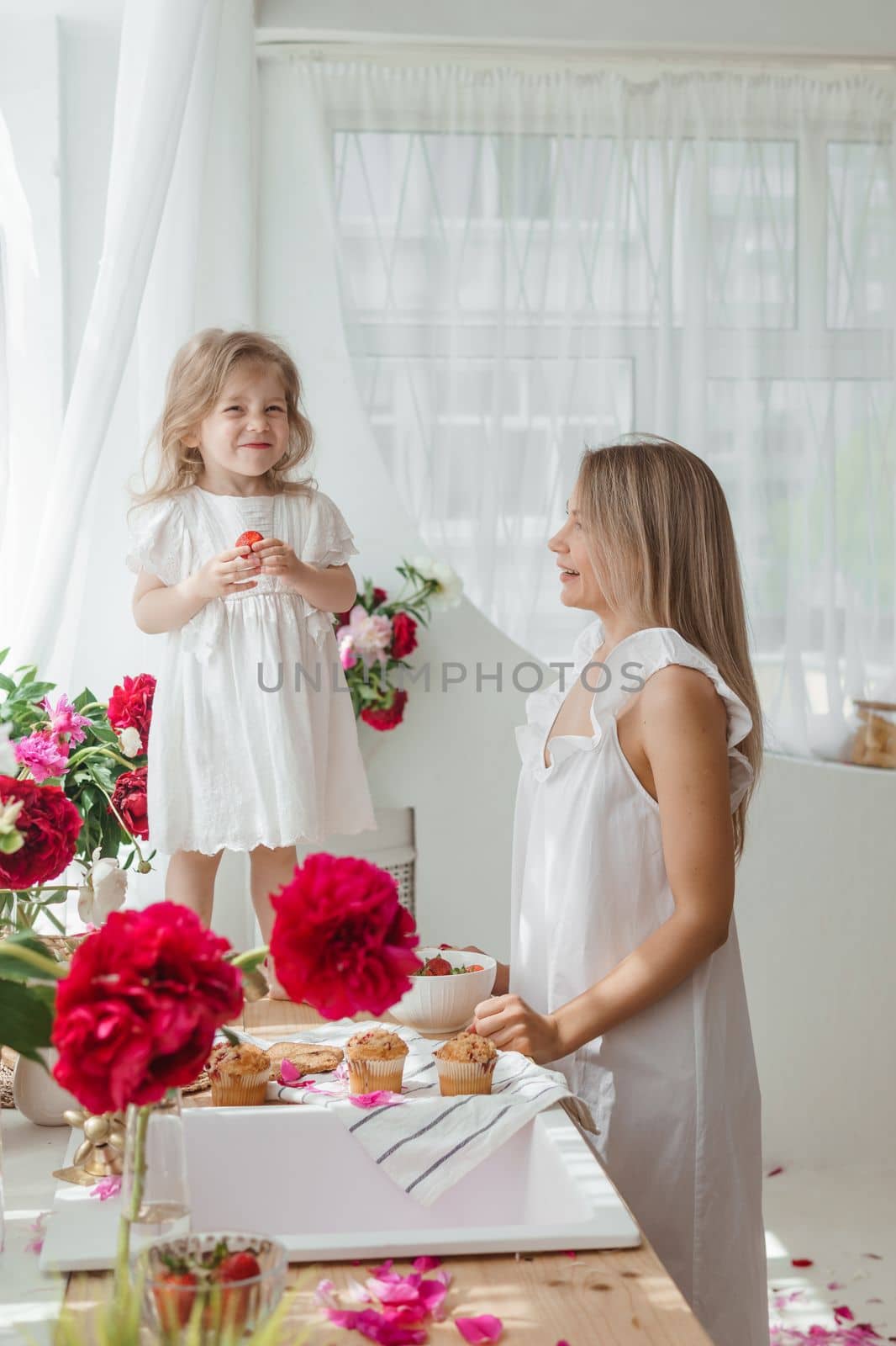 A little blonde girl with her mom on a kitchen countertop decorated with peonies. The concept of the relationship between mother and daughter. Spring atmosphere.