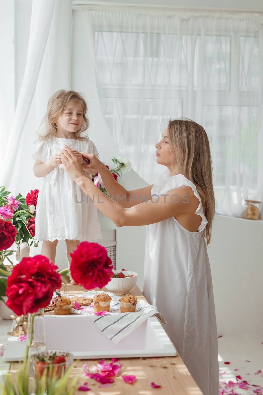 A little blonde girl with her mom on a kitchen countertop decorated with peonies. The concept of the relationship between mother and daughter. Spring atmosphere.