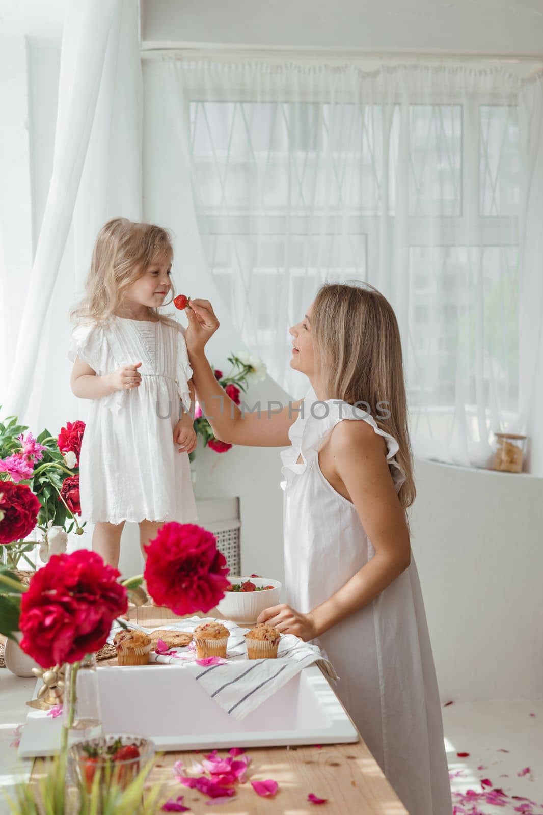 A little blonde girl with her mom on a kitchen countertop decorated with peonies. The concept of the relationship between mother and daughter. Spring atmosphere.