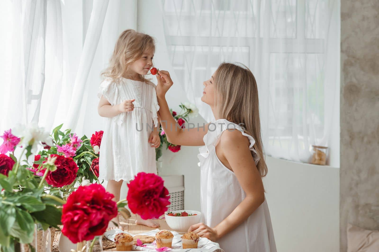 A little blonde girl with her mom on a kitchen countertop decorated with peonies. The concept of the relationship between mother and daughter. Spring atmosphere.