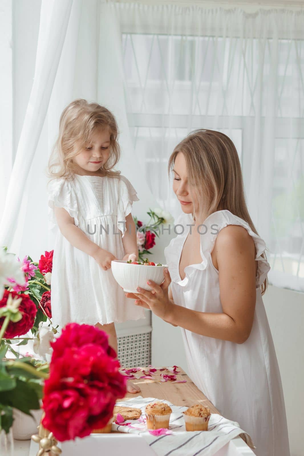 A little blonde girl with her mom on a kitchen countertop decorated with peonies. The concept of the relationship between mother and daughter. Spring atmosphere.