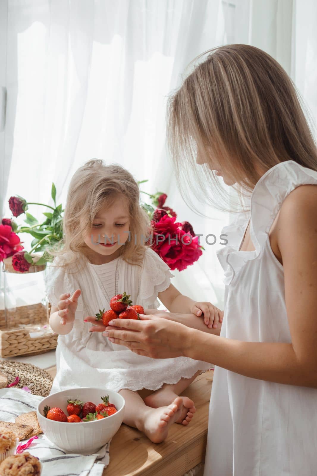 A little blonde girl with her mom on a kitchen countertop decorated with peonies. The concept of the relationship between mother and daughter. Spring atmosphere.