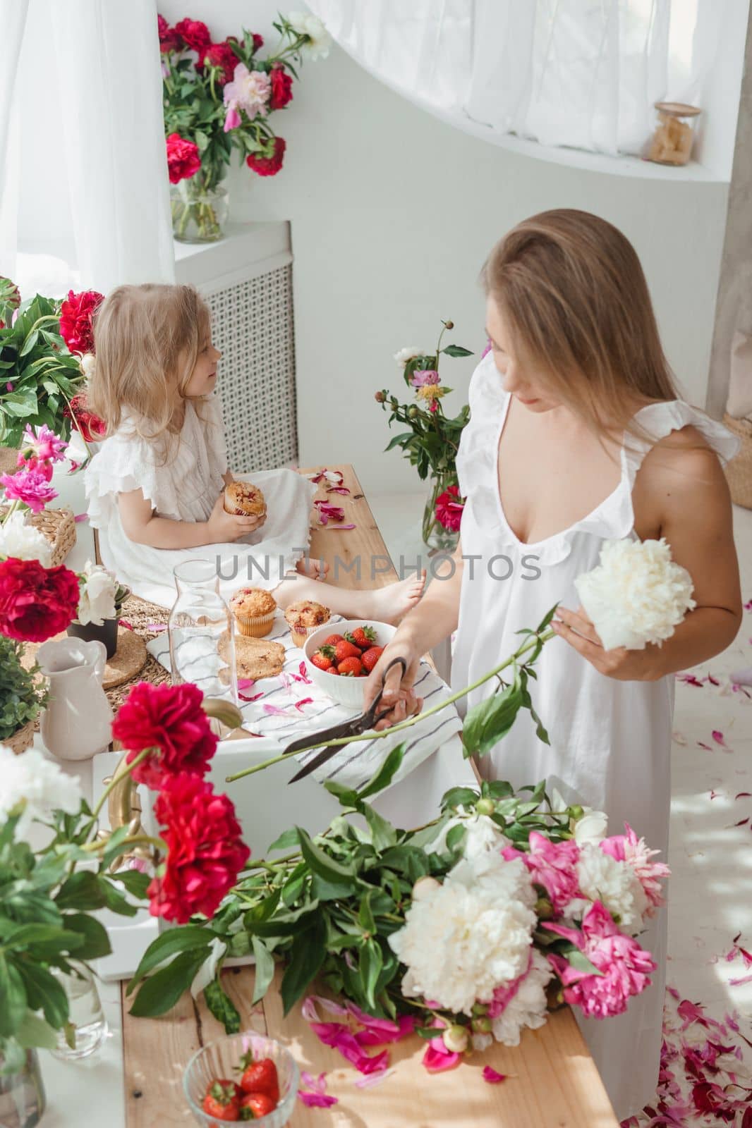 A little blonde girl with her mom on a kitchen countertop decorated with peonies. The concept of the relationship between mother and daughter. Spring atmosphere.