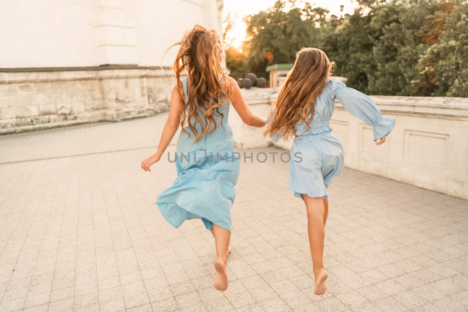 Daughter mother run holding hands. In blue dresses with flowing long hair against the backdrop of a sunset and a white building. by Matiunina