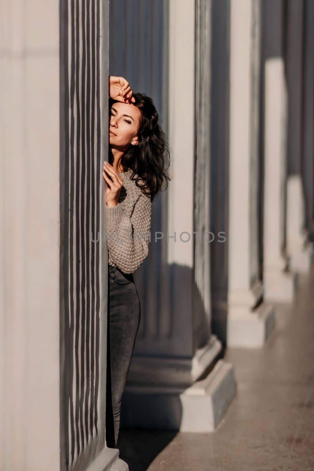 Woman building columns. An athletic woman in her 40s, dressed in a beige sweater and black jeans, poses near the pillars of a building. Walking around the city, tourism