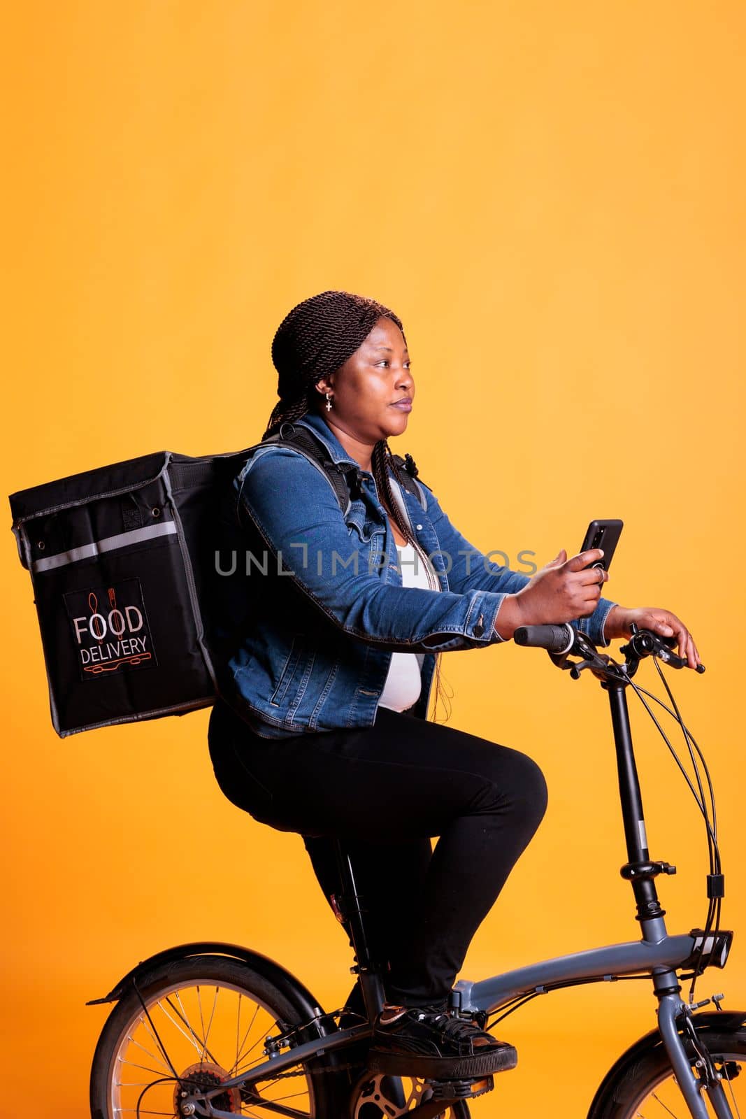 Pizzeria worker riding bike while delivering takeout food to client during lunch time, holding smartphone to check customer address on fast food app. Food transportation service and takeaway concept