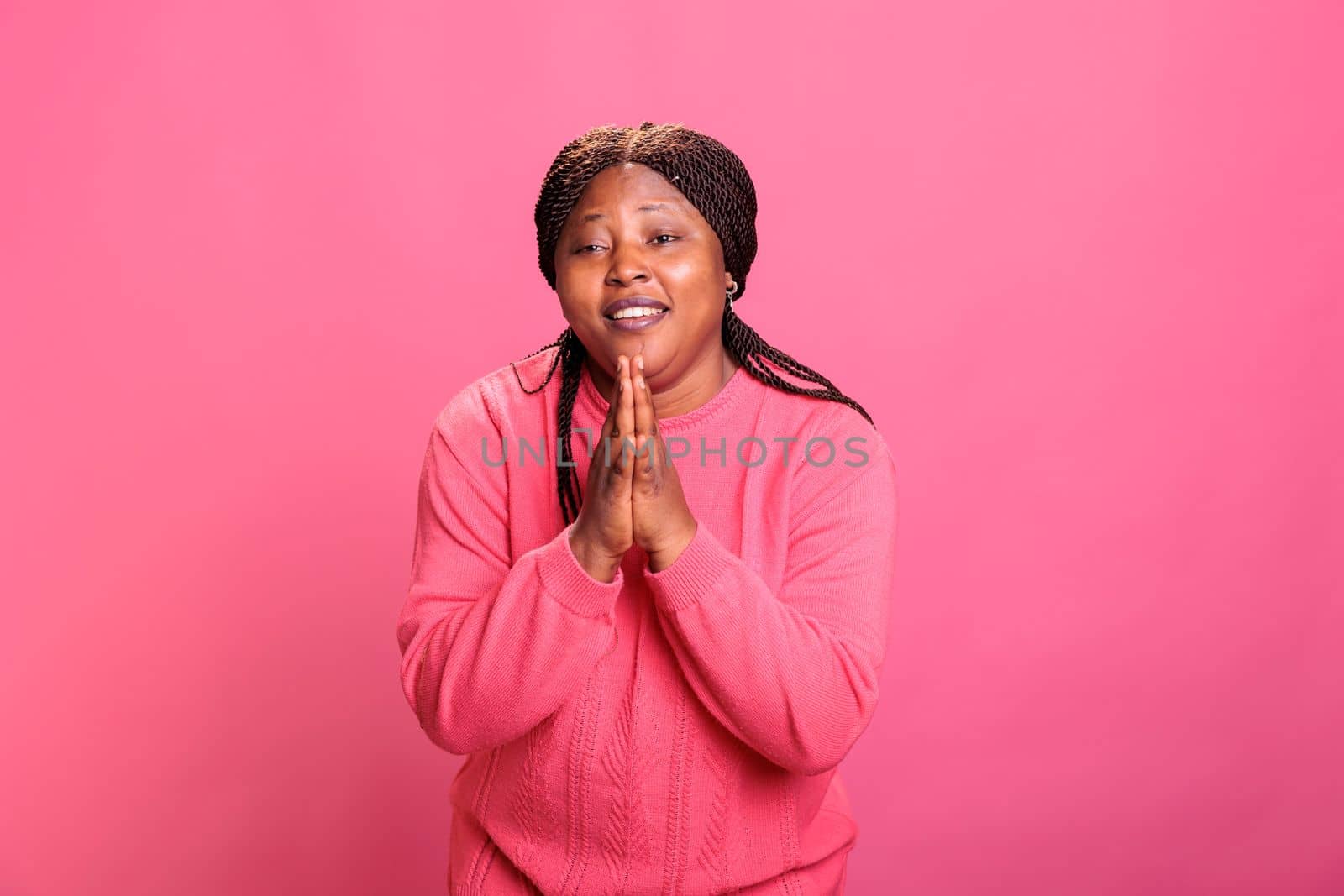 Happy female person doing prayer hands sign in studio, showing religious spiritual gesture over pink background. Young model with confidence praying to jesus for peace, christianity.