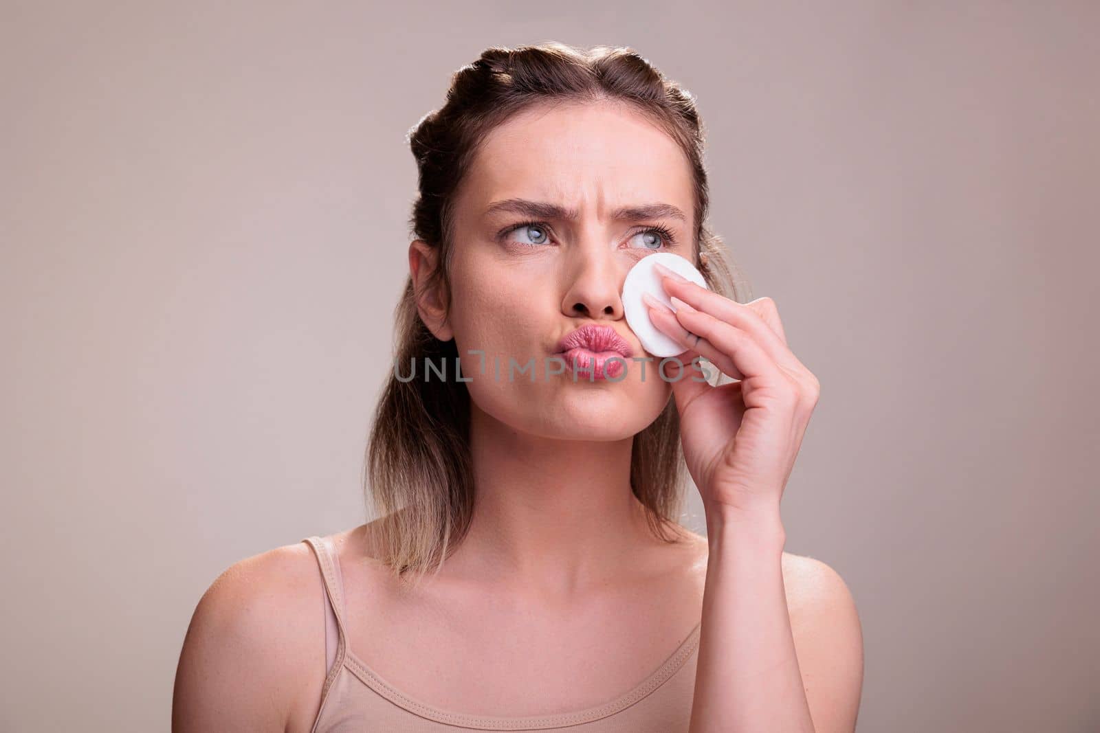 Skincare model cleansing face with cotton disk pad, doing skin treatment cleaning routine. Beautiful blonde young woman wearing beige top using micellar oil to remove make up