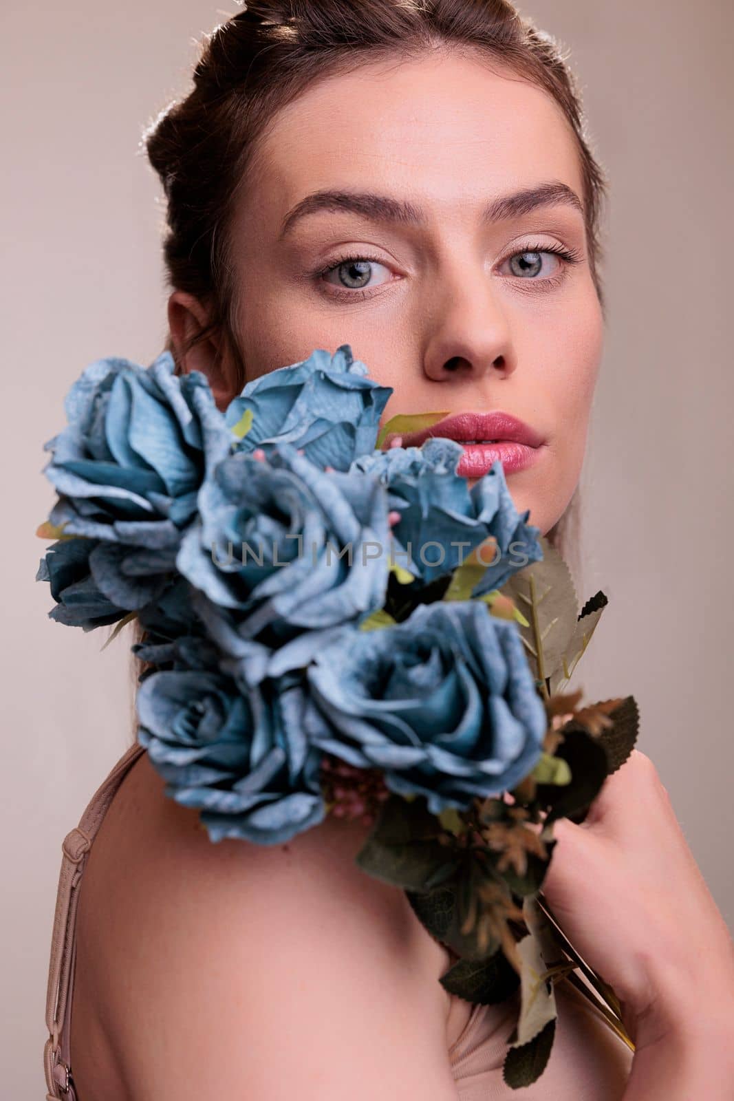 Beautiful woman posing with blue roses bunch on shoulder and looking at camera. Elegant young lady wearing natural make up holding blooming flowers romantic gift in arms close up portrait