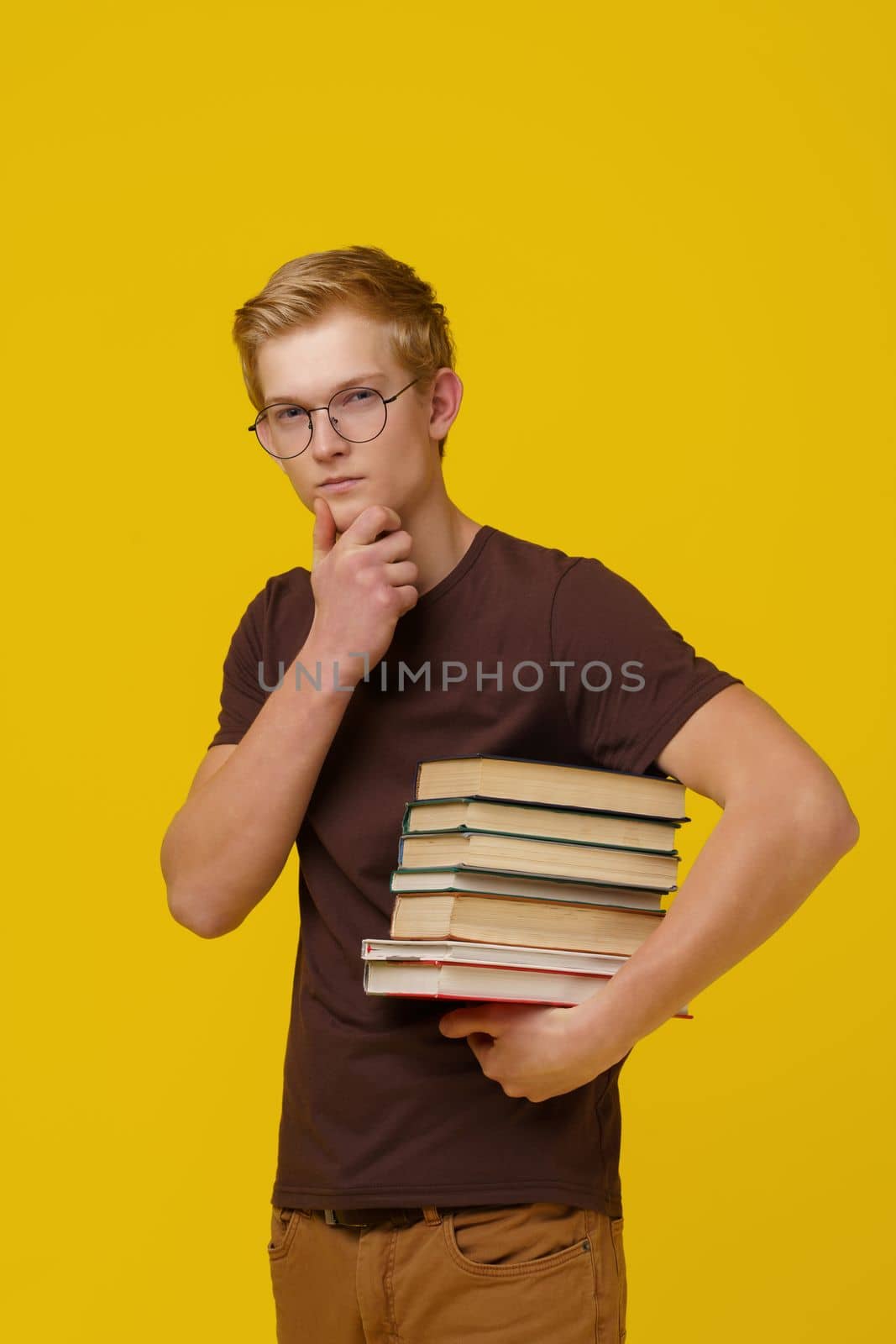 Knowledge Acquisition, Education, Concept Training. A Brooding Young European Man in a Brown T-Shirt Holds a Stack of Books in His Hand on a Yellow Background. High quality photo