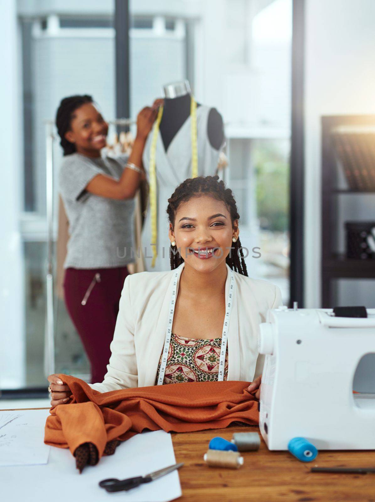 Working hard to achieve our goals. Portrait of a young fashion designer sewing garments while a colleague works on a mannequin in the background