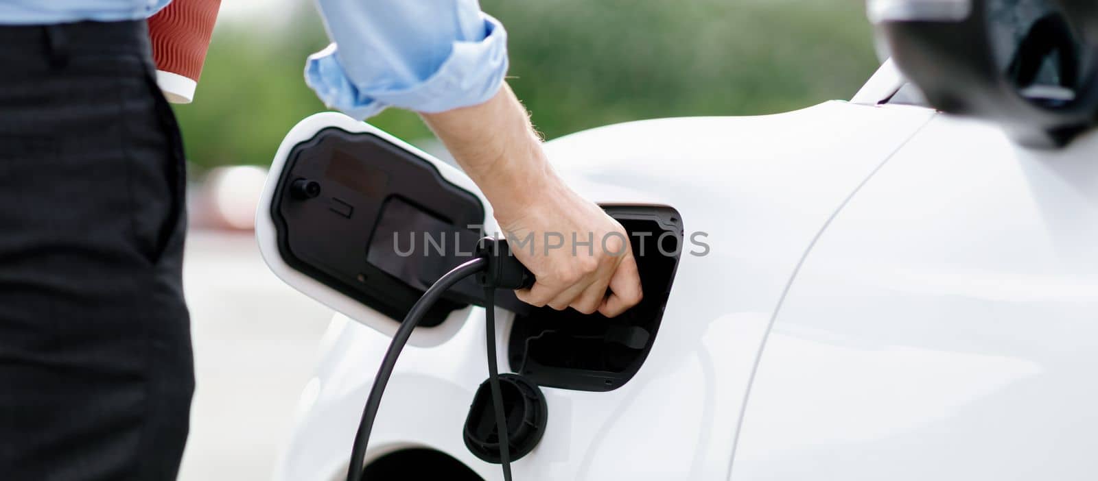 Closeup progressive businessman plugs charger plug from charging station to EV. by biancoblue