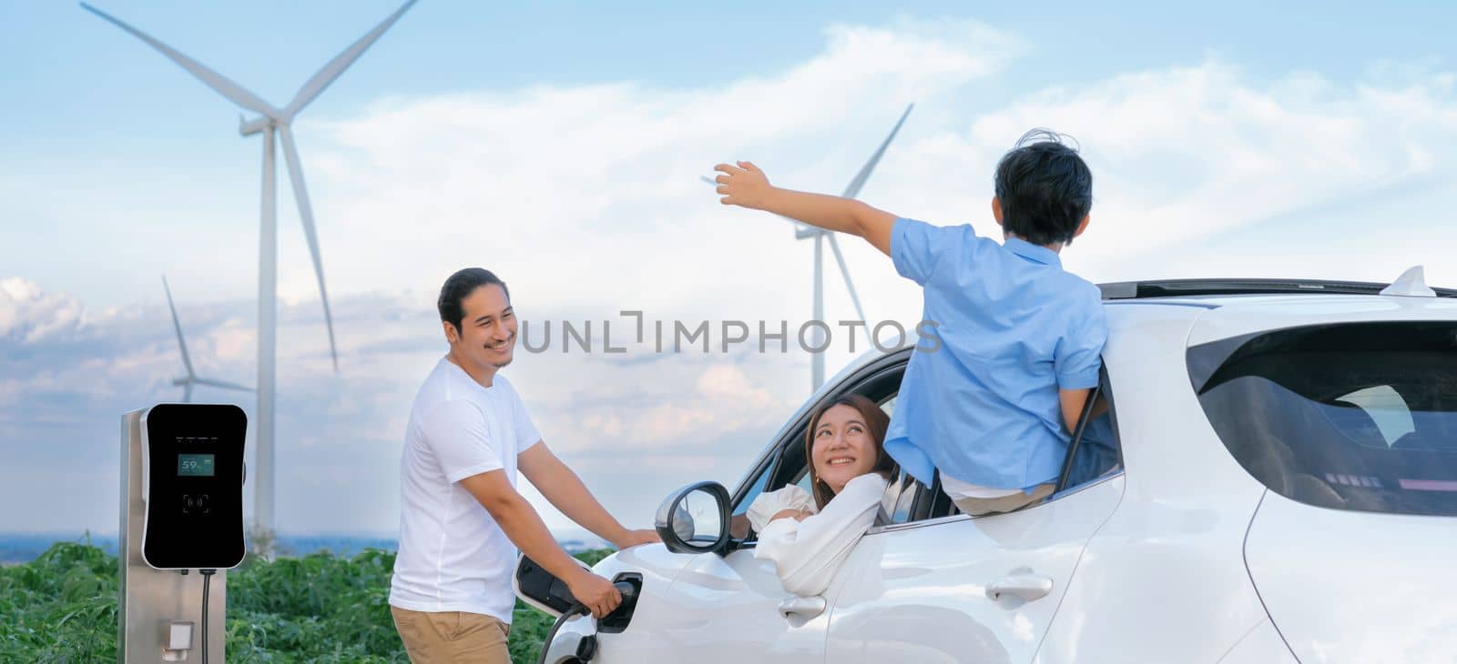 Concept of progressive happy family enjoying their time at wind farm with electric vehicle. Electric vehicle driven by clean renewable energy from wind turbine generator for charging station.