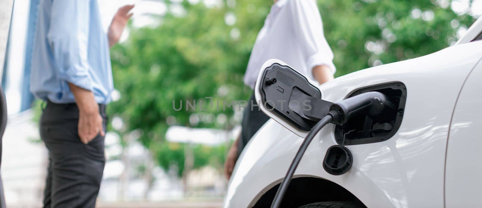 Focus parking-electric car connected to public charging station with blur progressive businesspeople holding coffee, residential building apartment and condo background for eco-friendly concept.