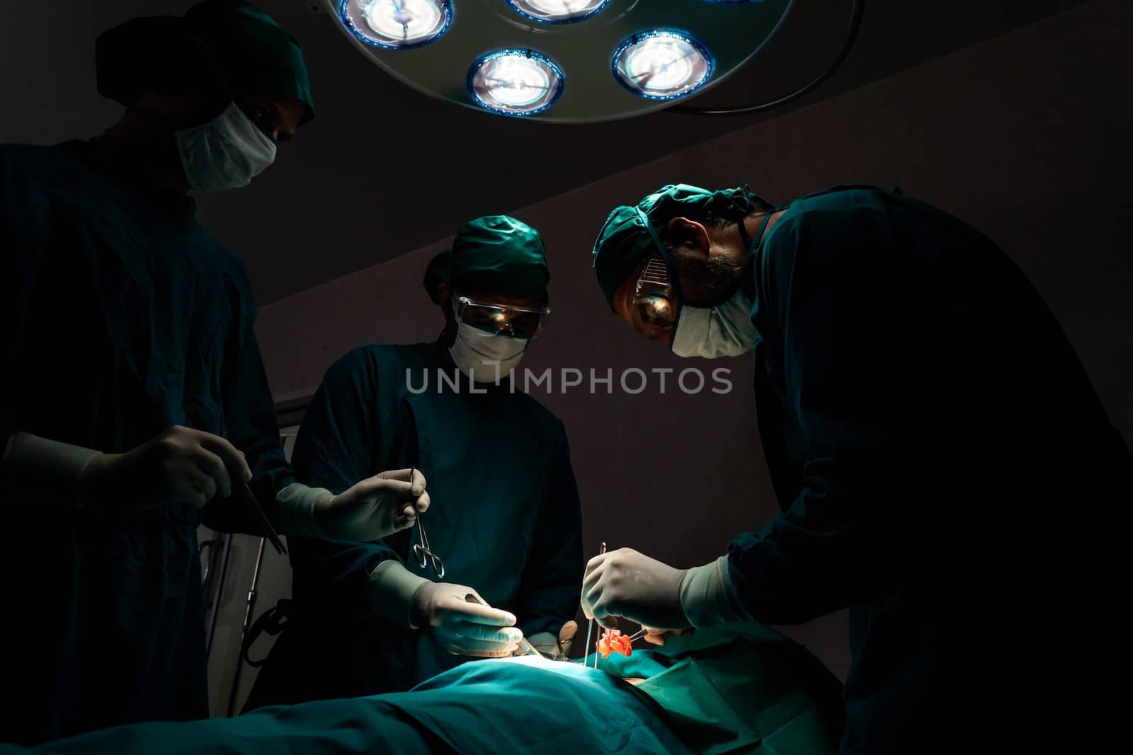 Surgical team performing surgery to patient in sterile operating room. In a surgery room lit by a lamp, a professional and confident surgical team provides medical care to an unconscious patient.