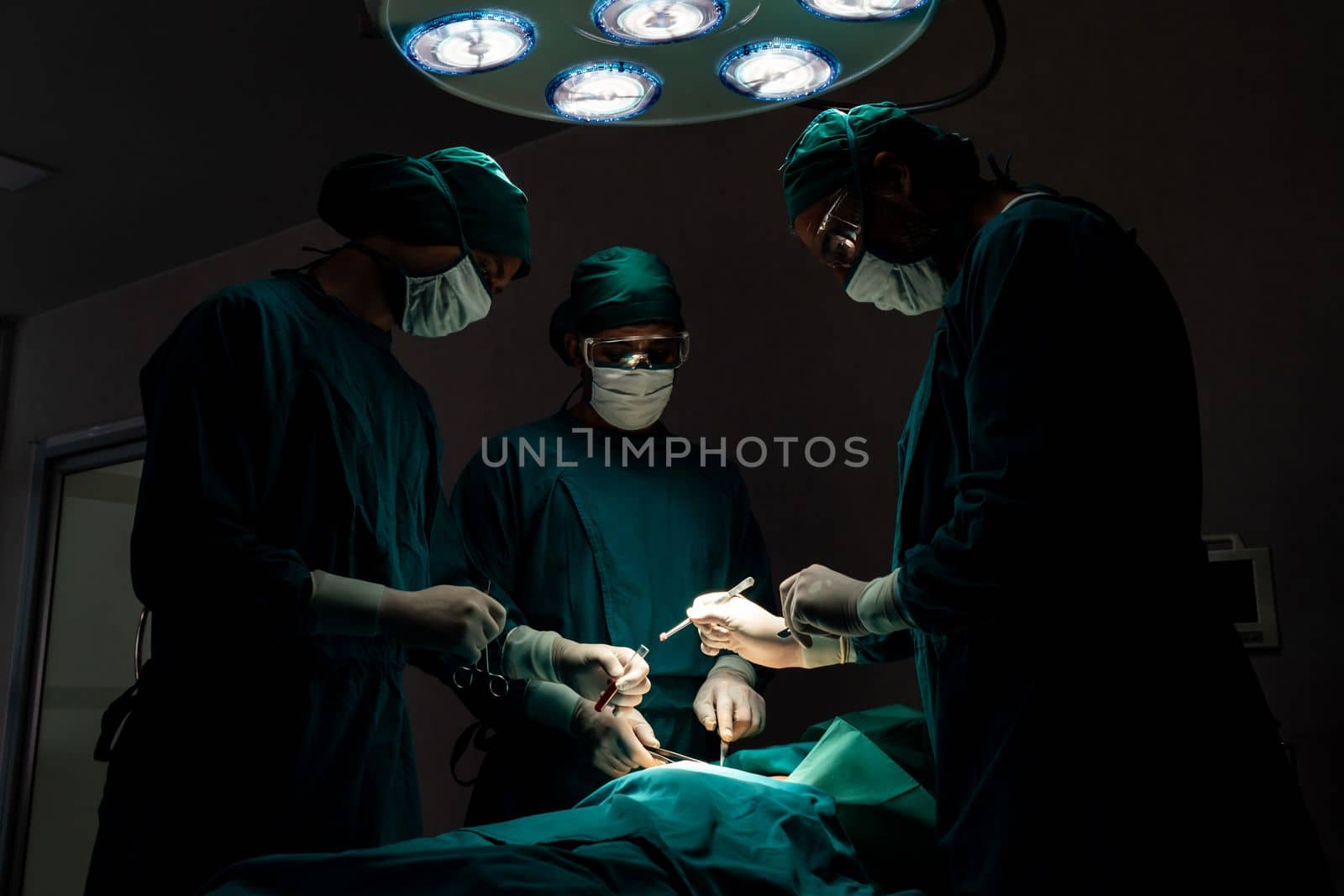 Surgical team performing surgery to patient in sterile operating room. In a surgery room lit by a lamp, a professional and confident surgical team provides medical care to an unconscious patient.