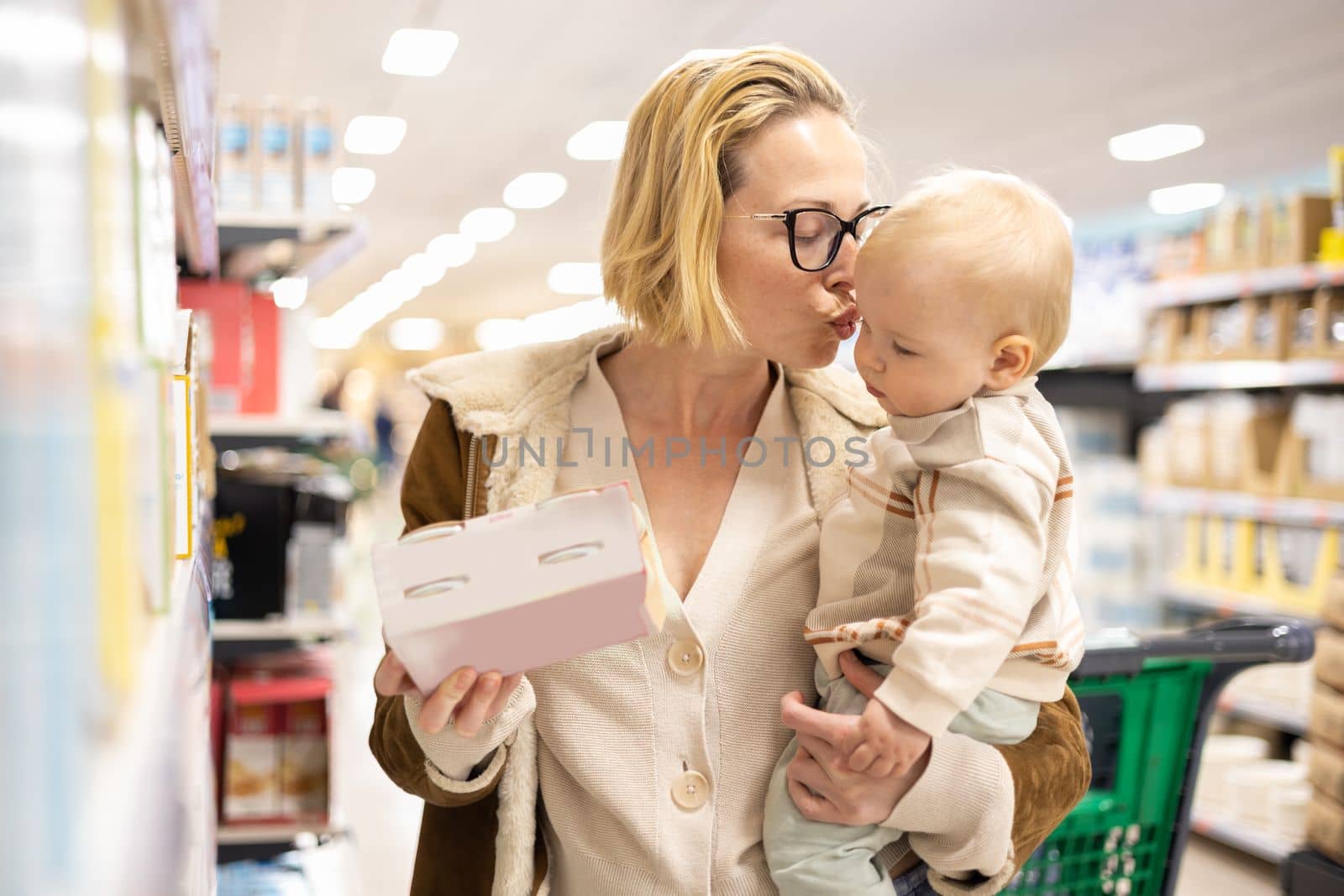 Caucasian mother shopping with her infant baby boy child choosing products in department of supermarket grocery store