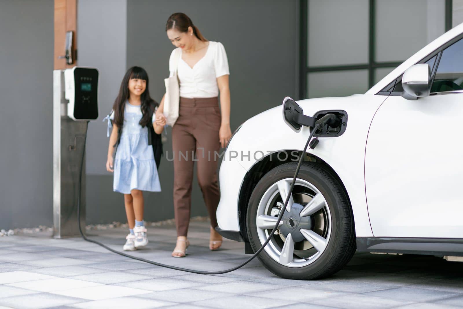 Focus EV car recharging at home charging station with blurred progressive woman and young girl in background for alternative clean energy technology concept for renewable electric vehicle.