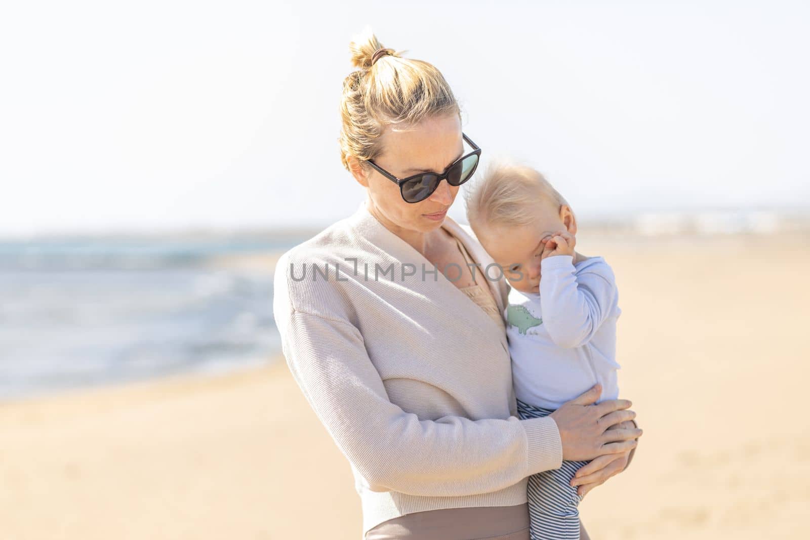 Mother holding and carrying his infant baby boy son on sandy beach enjoying summer vacationson on Lanzarote island, Spain. Family travel and vacations concept. by kasto