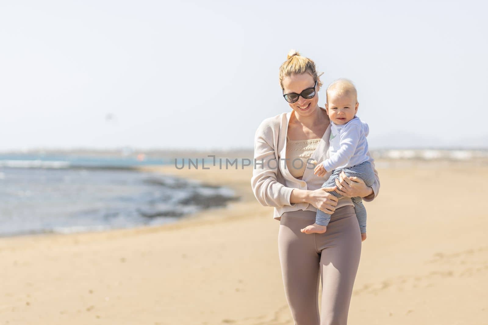 Mother holding and carrying his infant baby boy son on sandy beach enjoying summer vacationson on Lanzarote island, Spain. Family travel and vacations concept
