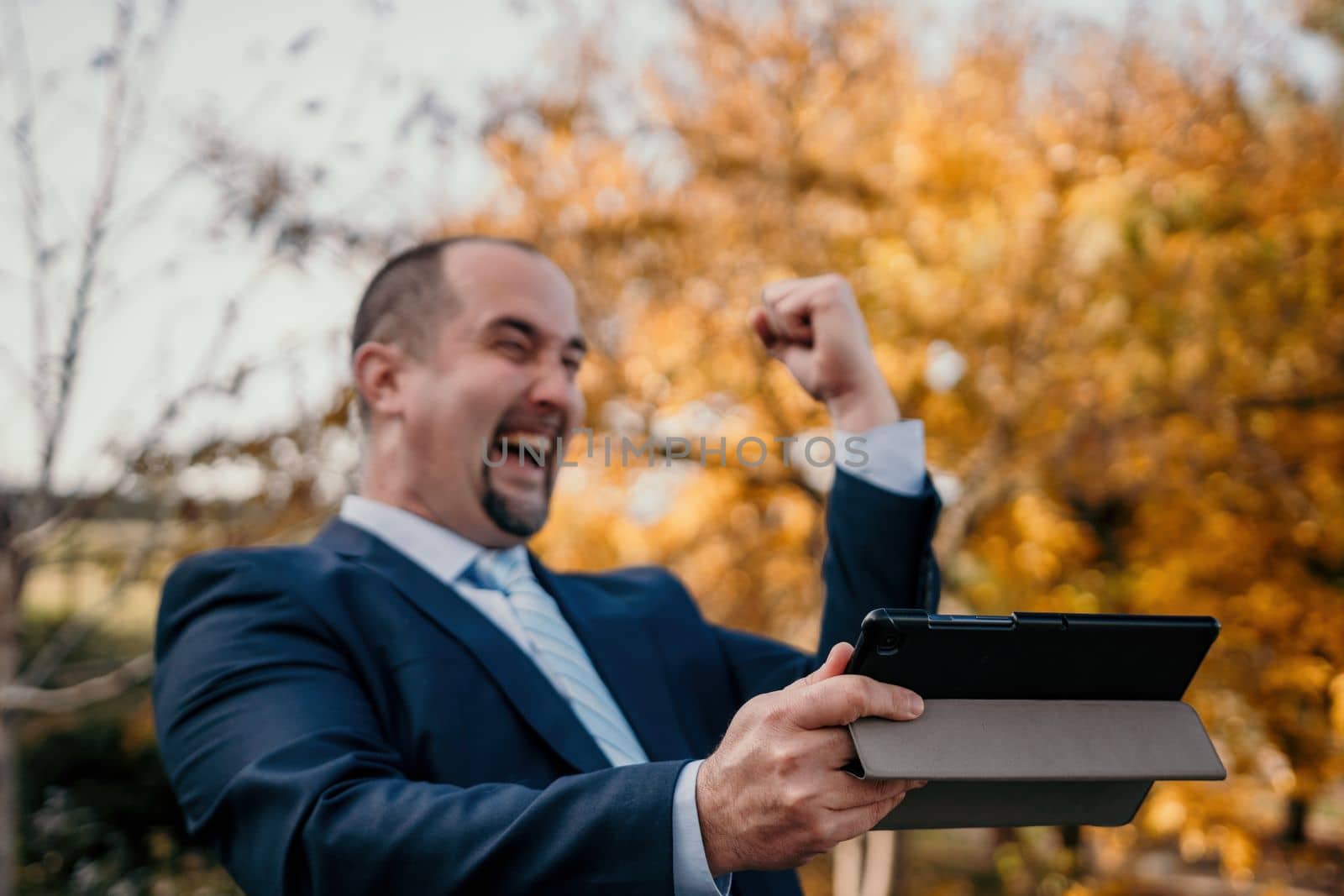 Abstrack defocused mature business man in suit reading a news sitting in the park on the bench. Entrepreneur drink coffee in autumn park. Senior executive rest in city park. by panophotograph