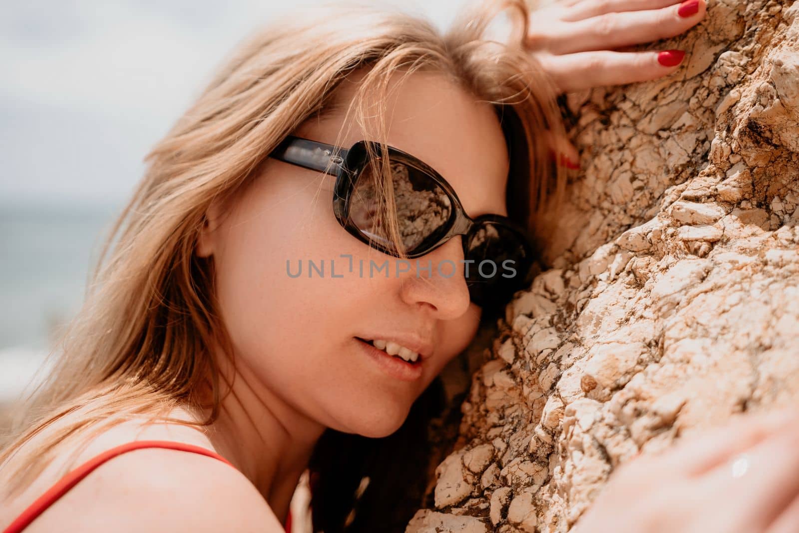 Young woman in red bikini on Beach. Blonde in sunglasses on pebble beach enjoying sun. Happy lady in one piece red swimsuit relaxing and sunbathing by turquoise sea ocean on hot summer day. Close up,