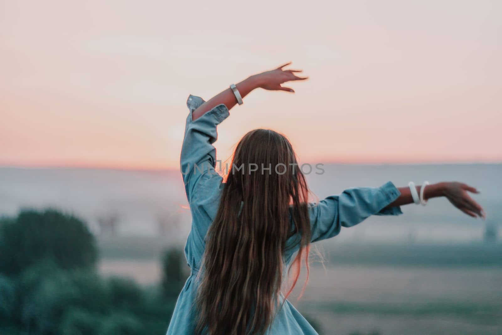 Happy woman standing with her back on the sunset in nature in summer with open hands. Romantic beautiful bride in white boho dress posing with mountains on sunset by panophotograph