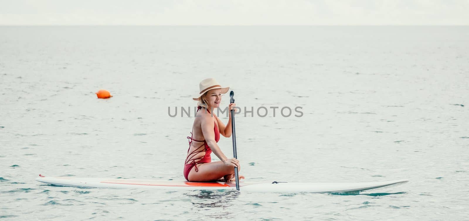 Woman in red bikini on sup board. Happy lady with blond hair in red bathing suit chilling and sunbathing by turquoise sea ocean on sunset. Holiday, vacation and recreational concept. by panophotograph