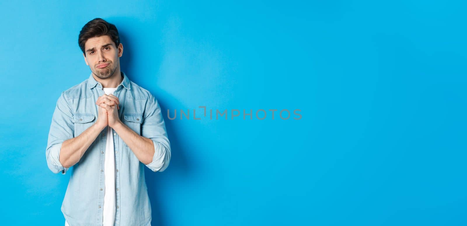 Young guy begging for help, pouting and looking sad while asking for favour, need something, standing against blue background.