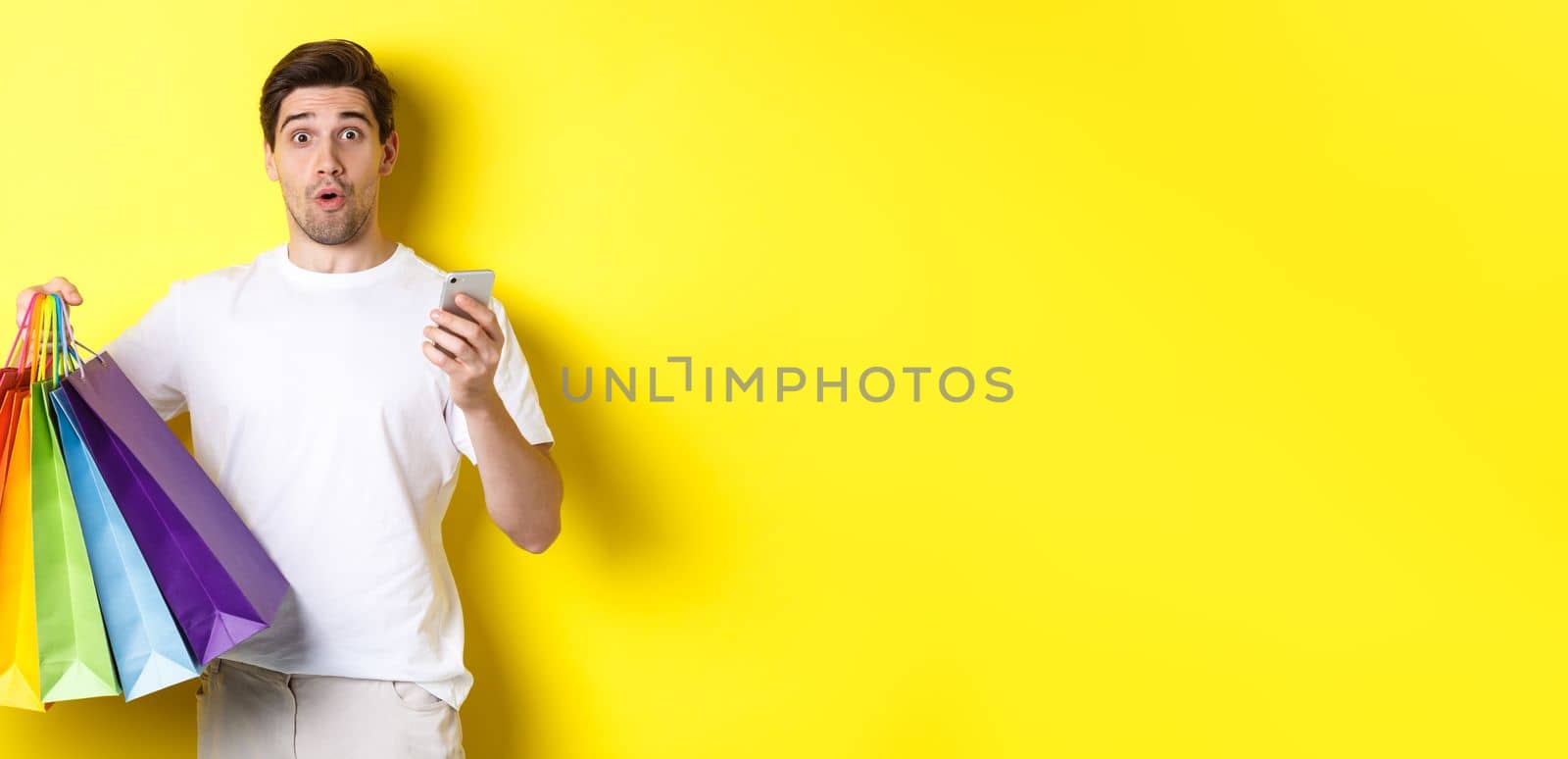 Concept of mobile banking and cashback. Surprised man holding shopping bags and smartphone, standing over yellow background.