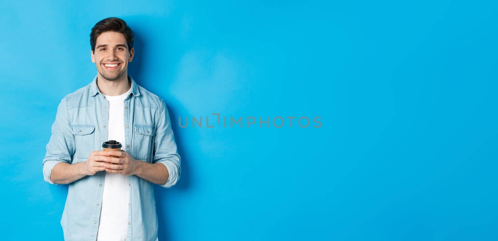 Young happy man drinking coffee from cafe takeaway, smiling pleased, standing against blue background.