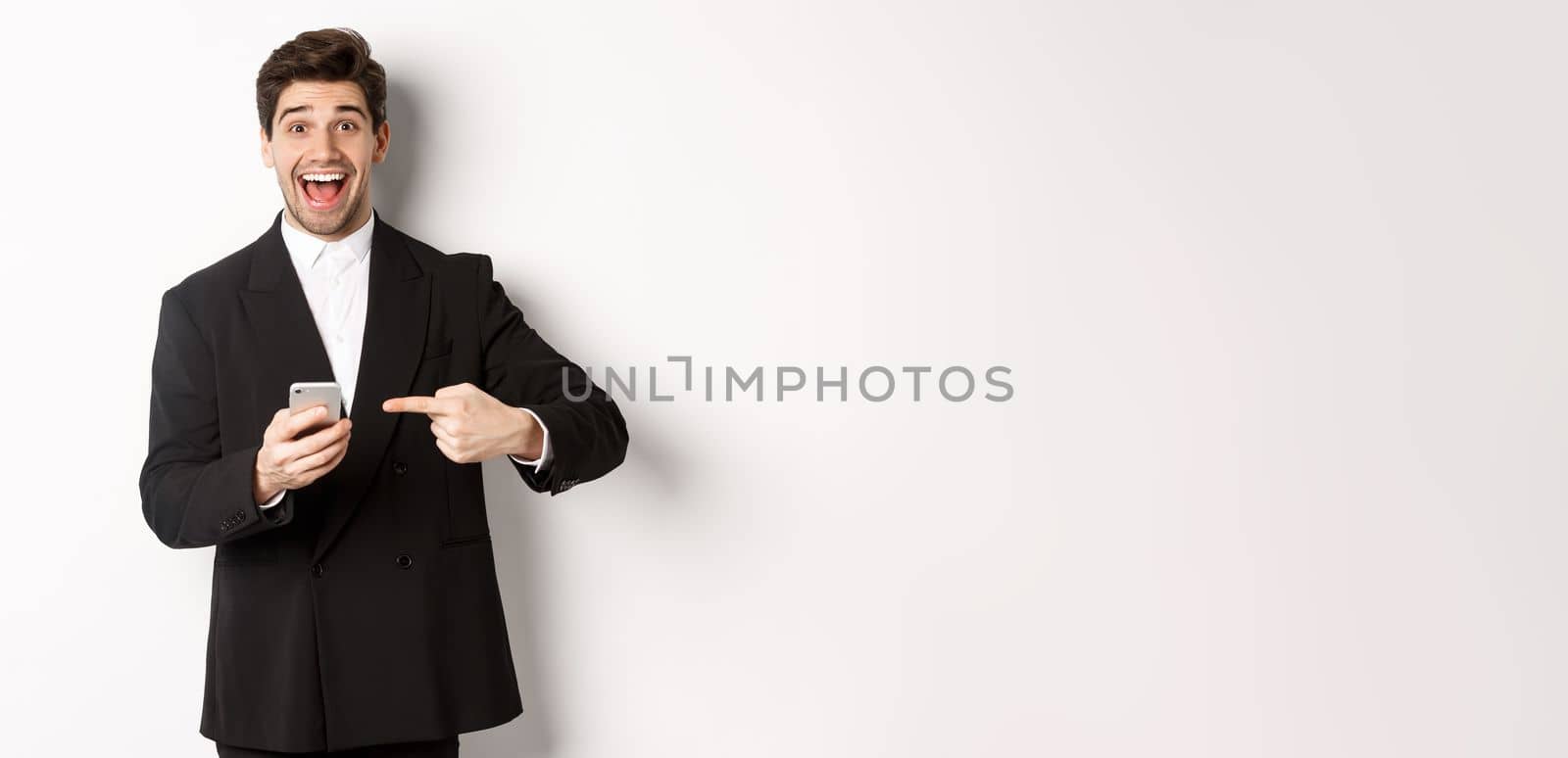 Image of cheerful businessman looking amazed, pointing at mobile phone, standing in suit over white background.