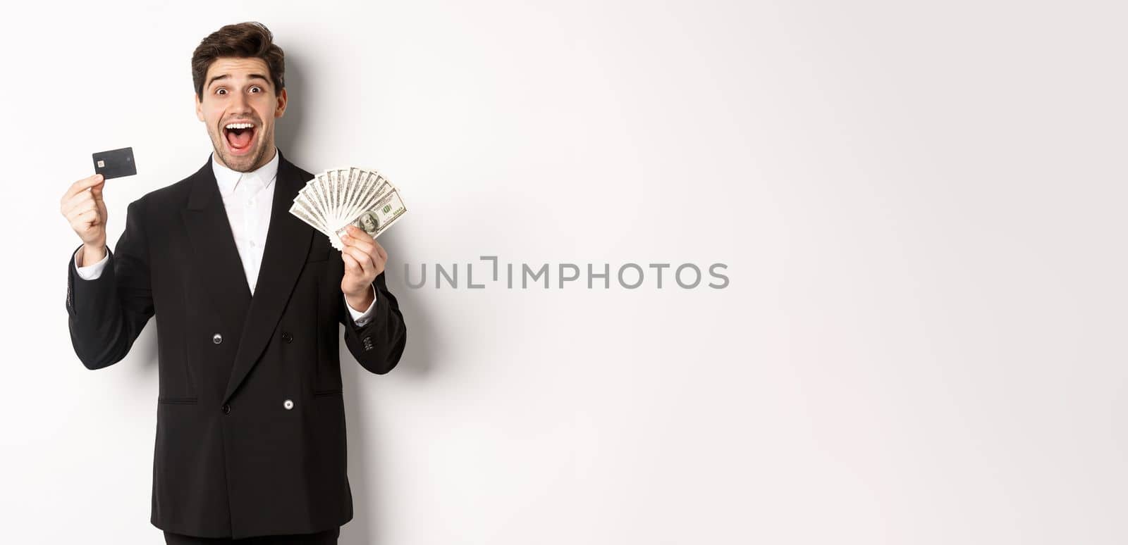 Image of handsome male entrepreneur in suit, showing credit card and money, smiling amazed, standing over white background.