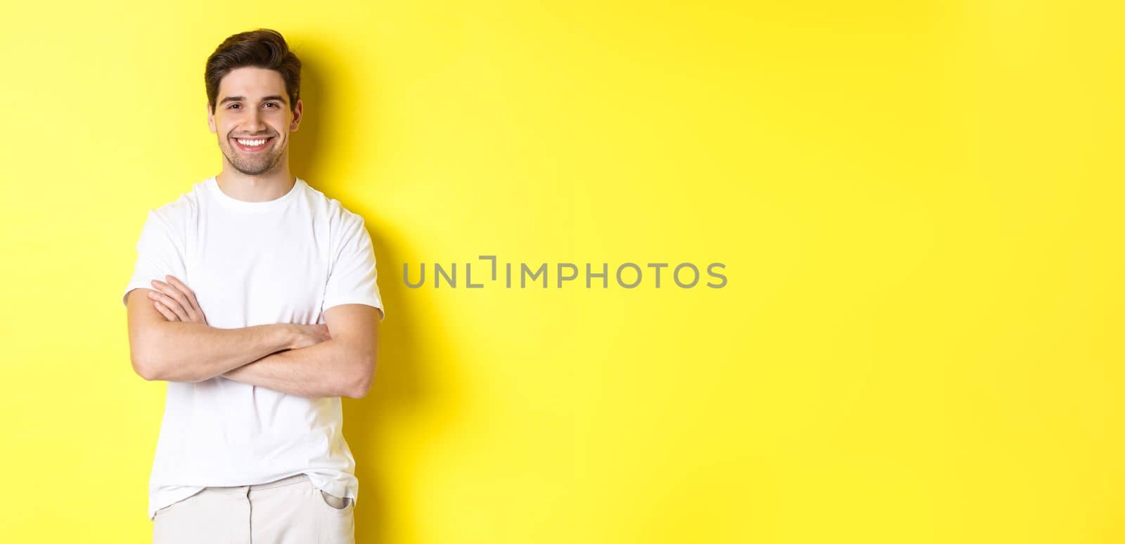 Image of confident caucasian man smiling pleased, holding hands crossed on chest and looking satisfied, standing over yellow background.